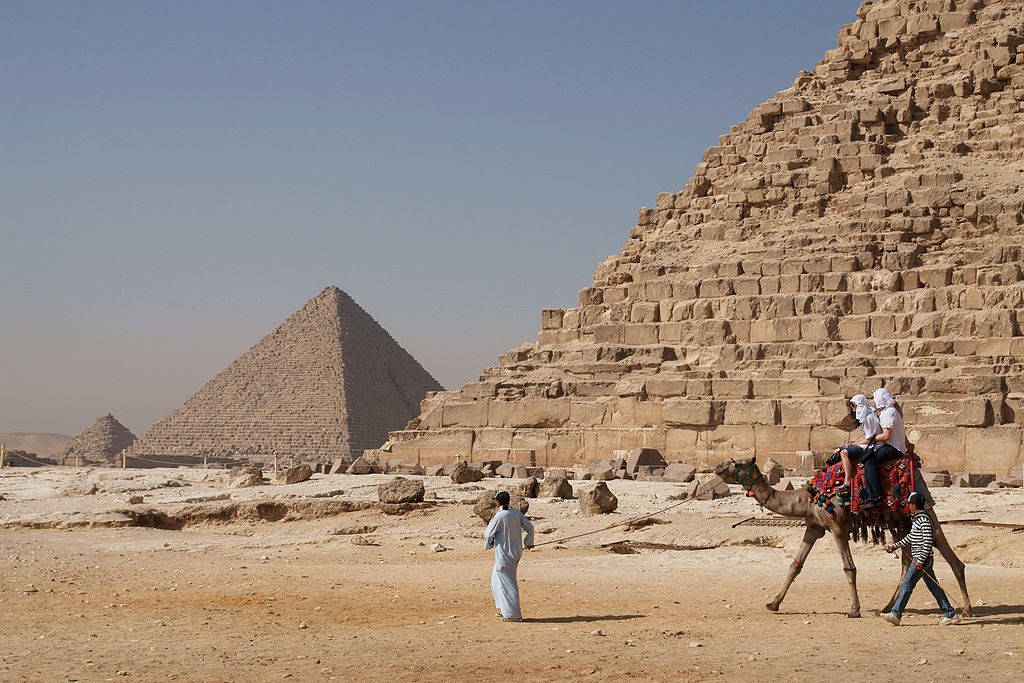 Tourists riding a camel in front of Pyramid of Khafre. The pyramid of Menkaure is in the background.